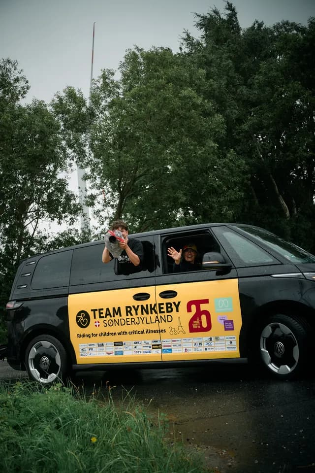 Helpers waving from a Team Rynkeby support car covered in sponsor logos